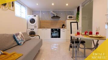 Un espacio habitable pequeño con sofá, una mesa de comedor para dos personas, lavadora y una kitchenette compacta.