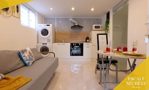 Un espacio habitable pequeño con sofá, una mesa de comedor para dos personas, lavadora y una kitchenette compacta.