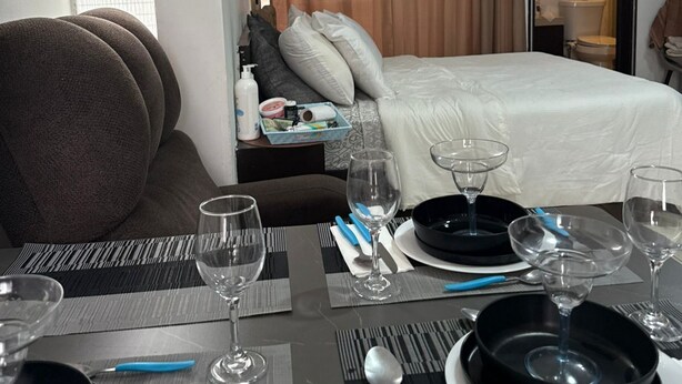 Ein Hotelzimmer mit einem ordentlich bezogenem Bett, einem für zwei Personen gedecktem Esstisch und einem Fenster mit Jalousien.