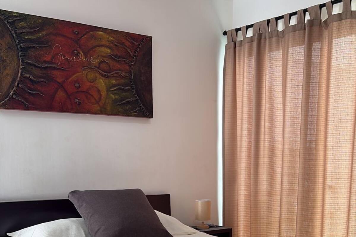 A neatly made bed with a white bedspread and two pillows, a painting on the wall, and a window with curtains.