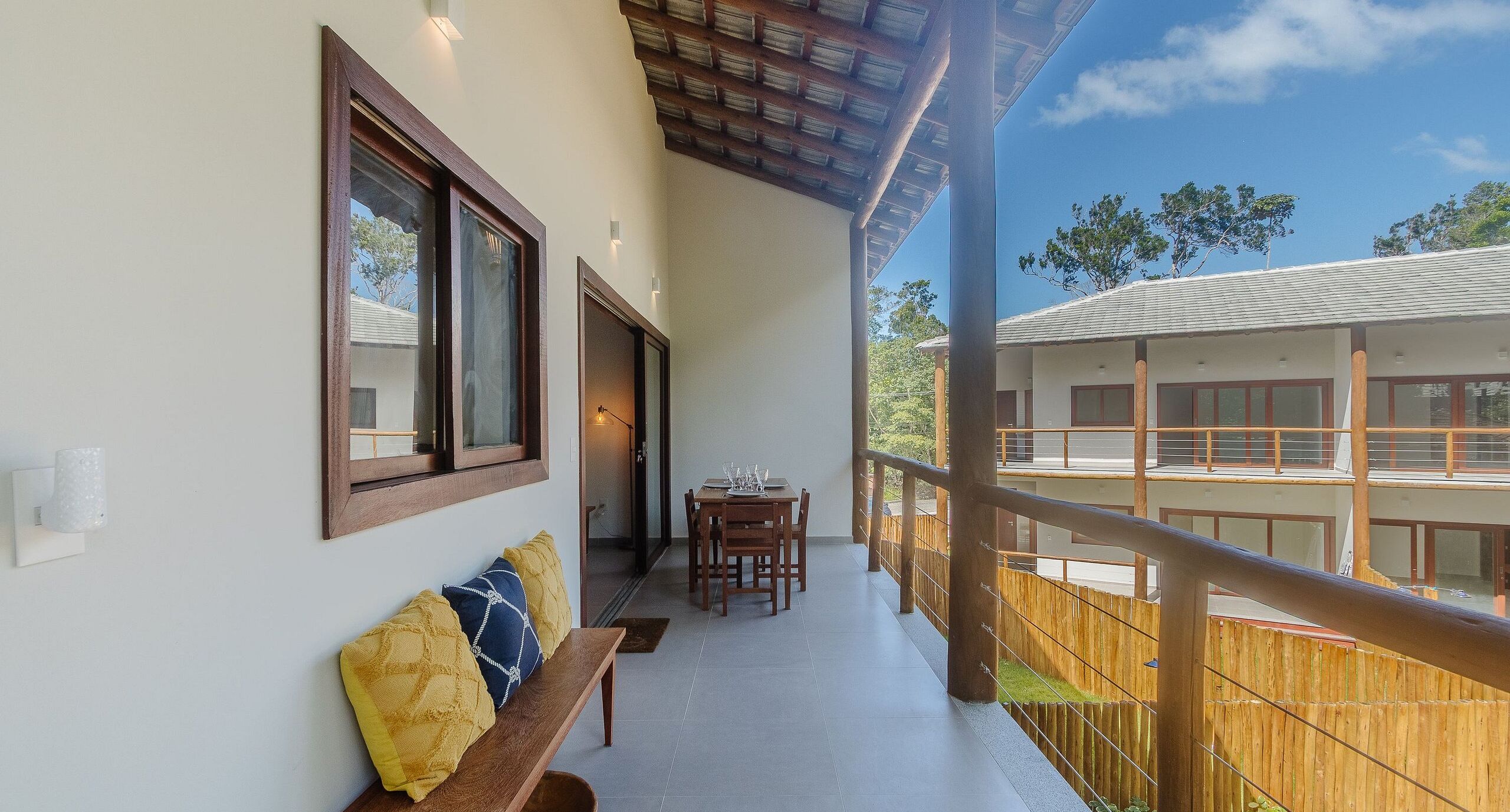 A balcony with a wooden bench and cushions, a dining table with chairs, and a view of another building.