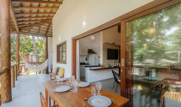 A dining area with a wooden table set for four, a hammock, and a kitchen visible through an open doorway. - Houses in the Ecovillage D'ajuda Condominium - EVL (Porto Seguro)