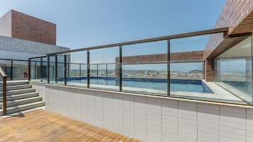 A rooftop swimming pool with glass railings, a wooden deck, and a city view.