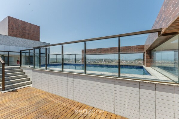 Una piscina en la azotea con barandillas de vidrio, terraza de madera y vista a la ciudad.
