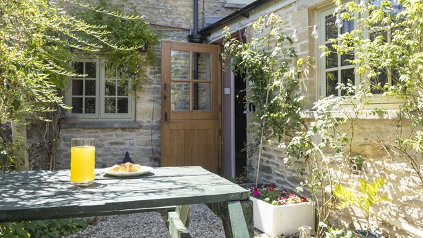 A stone building with a wooden door and windows, a table with a glass of orange juice and a plate of food, and potted plants. - The Rabbit Hole | Cotswold Holiday Let | 1 Bed & Sleeps 2 (Langford, Lechlade)