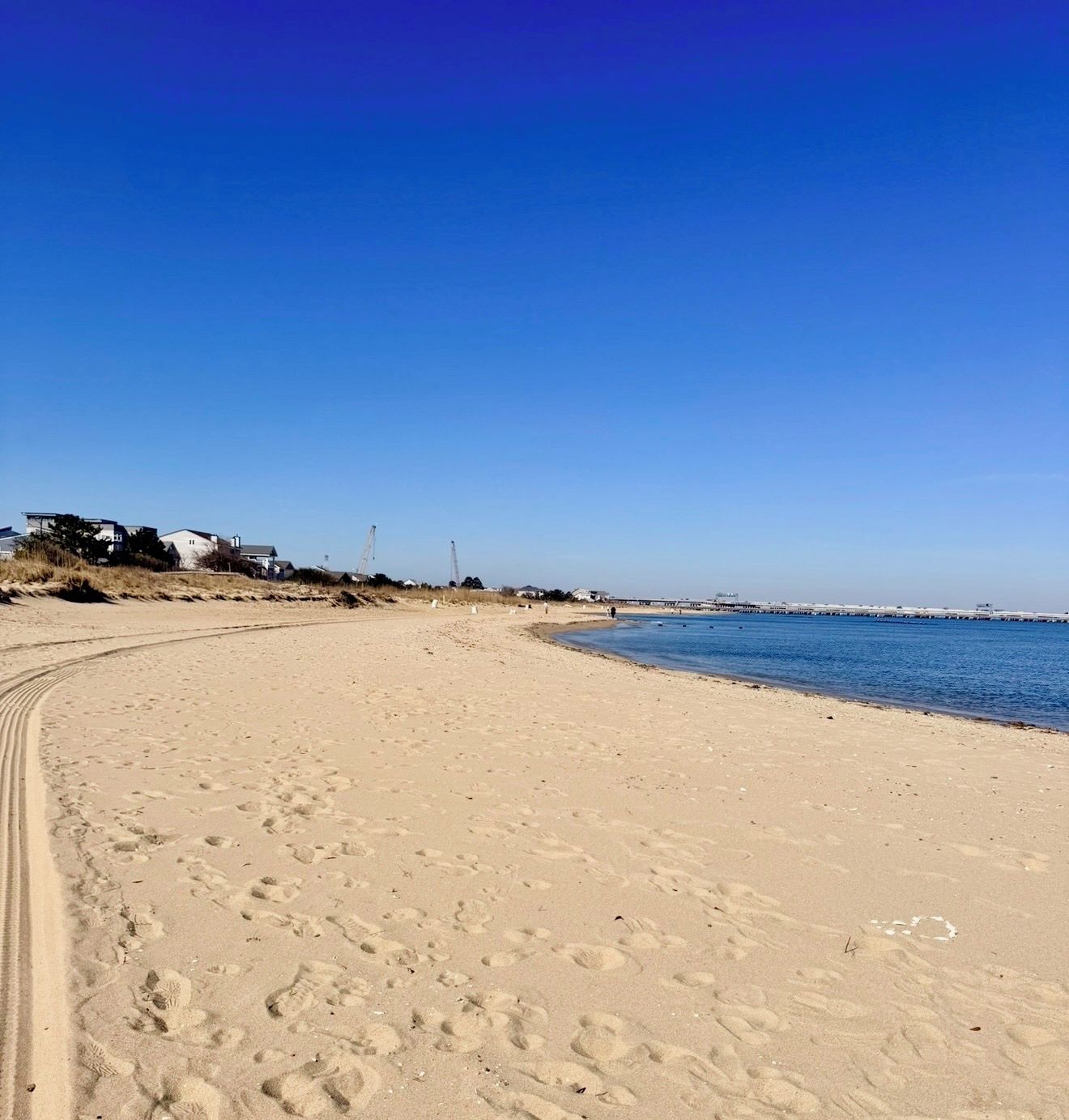 A sandy beach with footprints, a clear blue sky, and a distant pier.