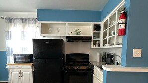 A compact kitchen with a black refrigerator, stove, and microwave. It features white cabinetry, a blue accent wall, and a window with curtains.
