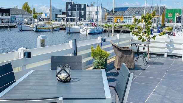En terrass med utsikt över vattnet, med ett bord och stolar, en krukväxt och en vy över båtar och byggnader.
