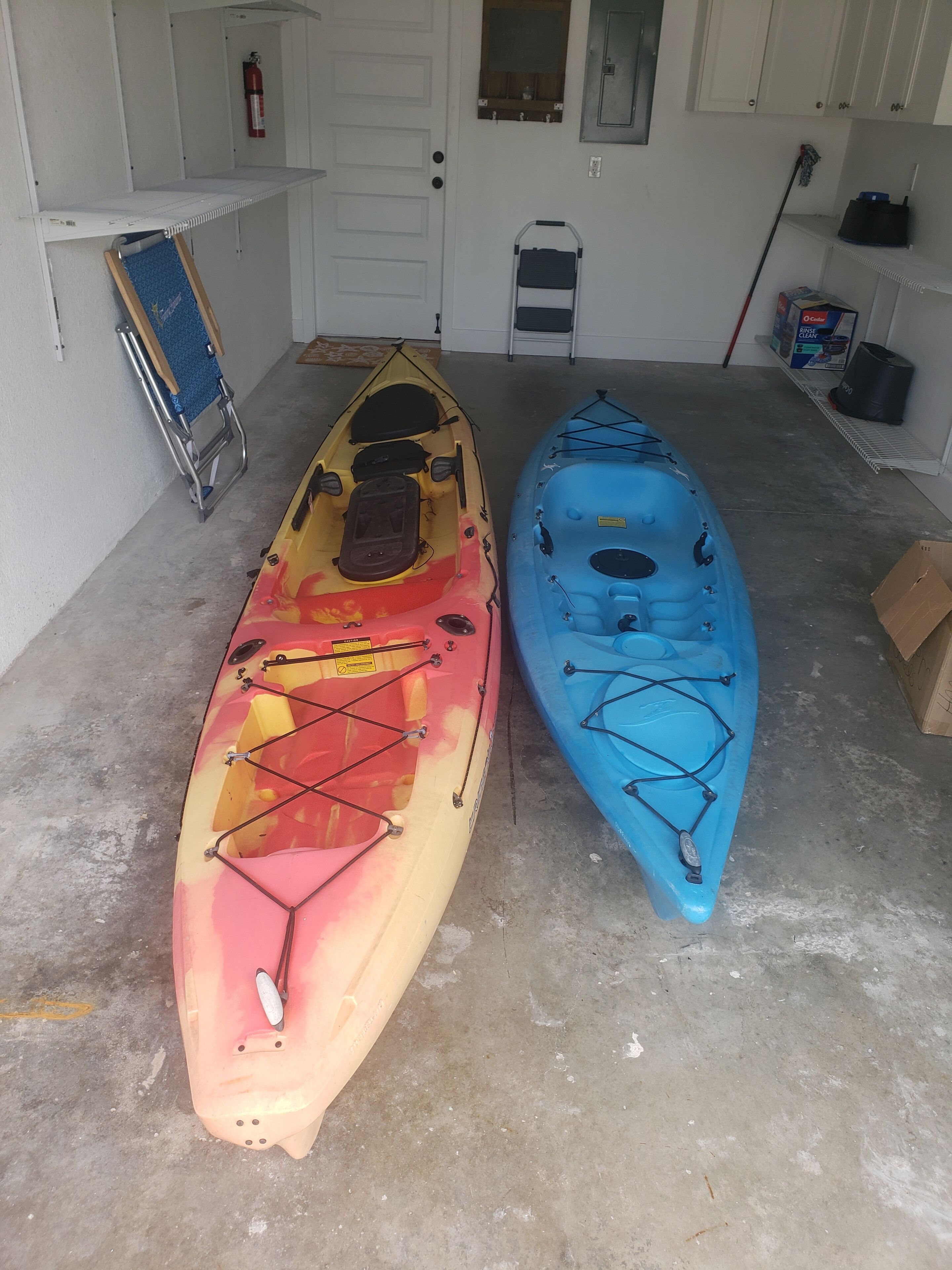 Two kayaks in a garage with a fire extinguisher, a ladder, and a broom.