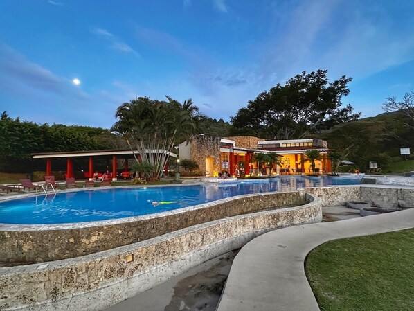 A pool area with a stone border, a covered seating area, and a building with red pillars.
