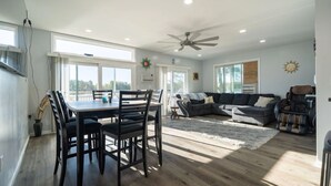 A spacious living room with a dining area, a ceiling fan, and large windows offering a view of the outdoors.