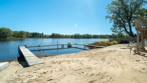 A sandy beach with a wooden dock extending into a calm lake, surrounded by trees and a wooden structure.