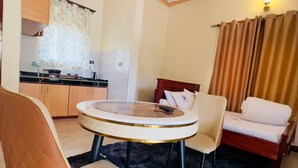 Een compacte hotelkamer met kitchenette, een rond dining table, twee stoelen, een bed en een groot raam met gordijnen.