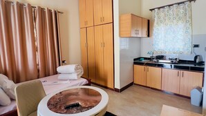Een compacte hotelkamer met een bed, een klein rond tafeltje, een stoel, een bank, een kitchenette en houten kastjes.