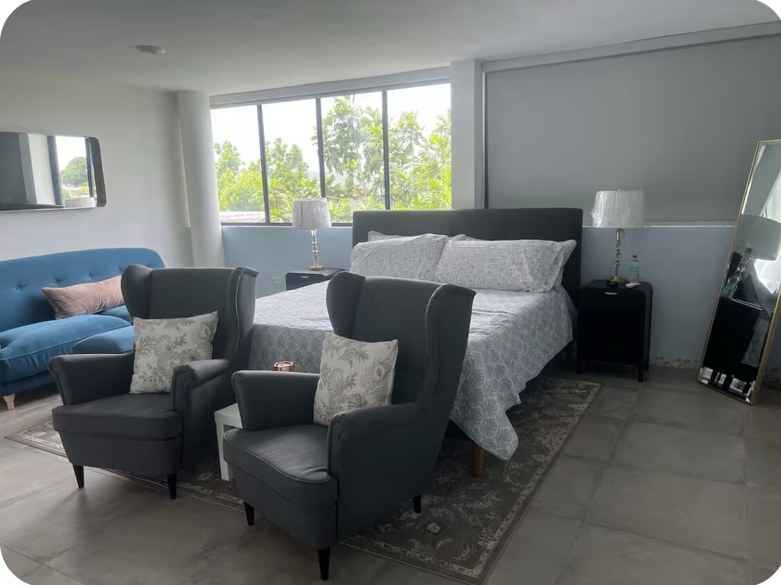Une chambre moderne avec un grand lit, deux fauteuils, un canapé bleu et une vue sur les palmiers par la fenêtre.