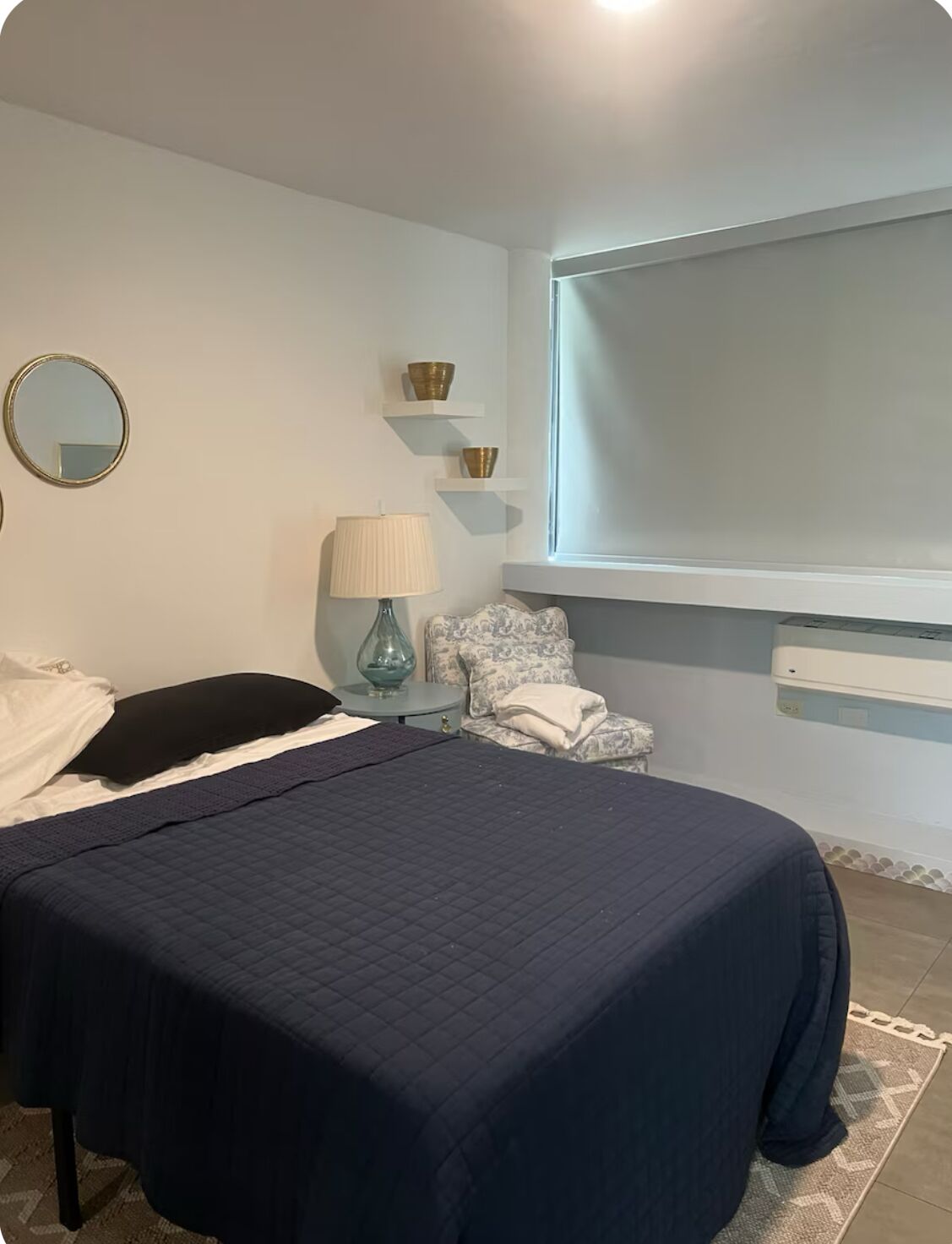 Un dormitorio con cama, lámpara de noche, espejo y ventana.