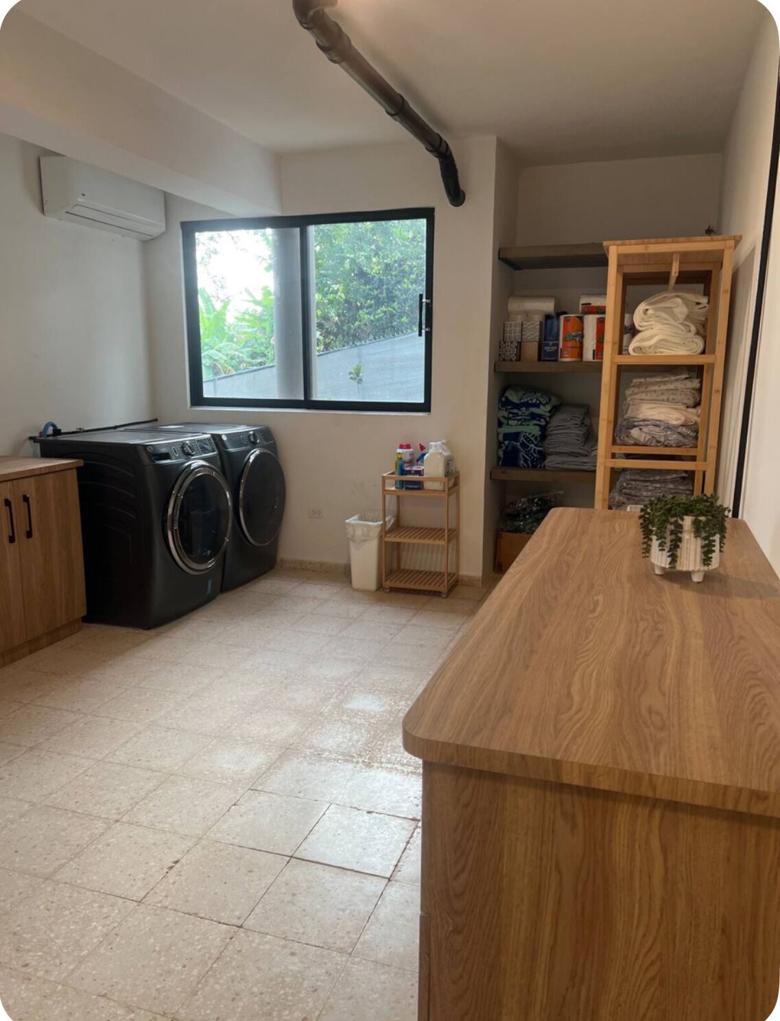 A laundry room with a washing machine, a drying rack, a small shelf with cleaning supplies, and a window with a view of greenery.