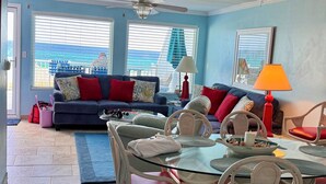 A living room with a blue sofa, red cushions, a glass coffee table, and white chairs. There are two windows with blinds, a ceiling fan, and a lamp on a side table.