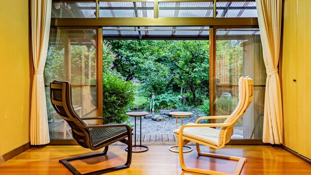 Una habitación con dos sillas y una mesita pequeña, ventanas amplias con cortinas y vista a un jardín.