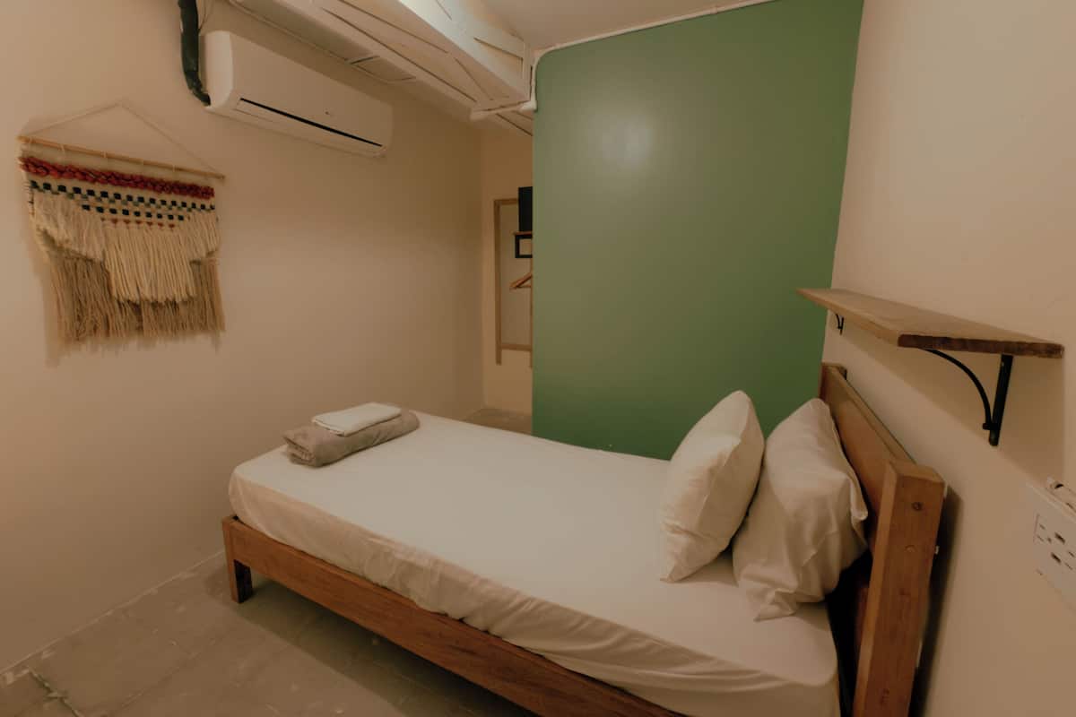 Quarto individual com cama de estrutura de madeira, parede verde e prateleira e ar condicionado fixados na parede.