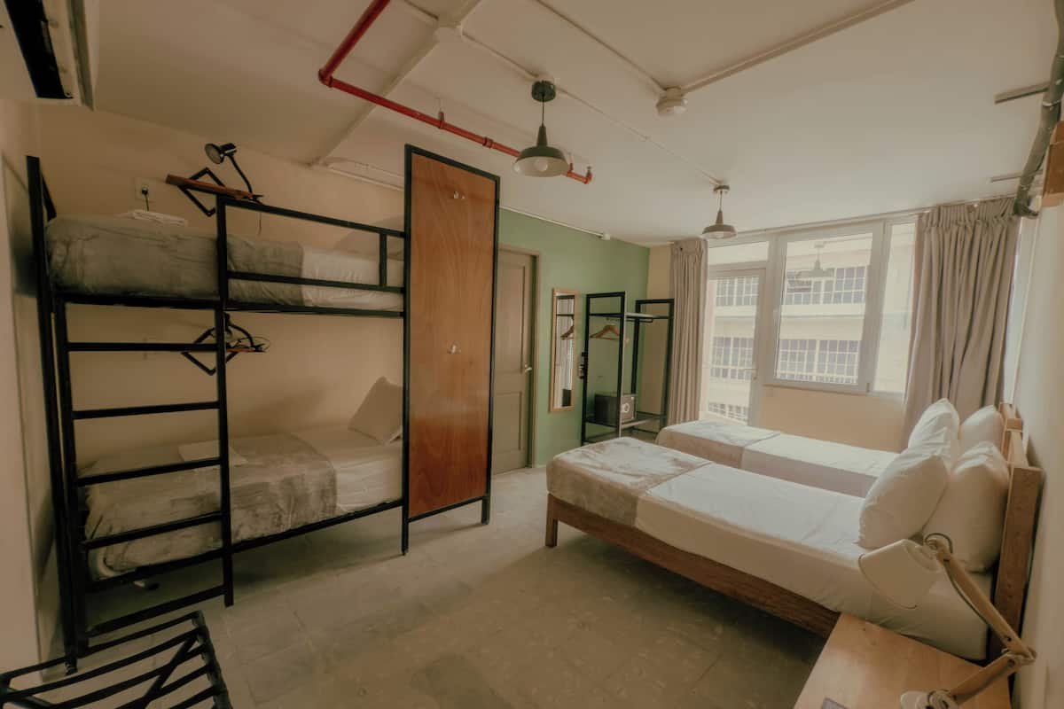 Um quarto de estudante com beliches, uma escrivaninha com luminária, um guarda-roupa e uma janela com cortinas.