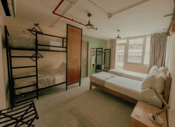 A dormitory room with bunk beds, a desk with a lamp, a wardrobe, and a window with curtains.