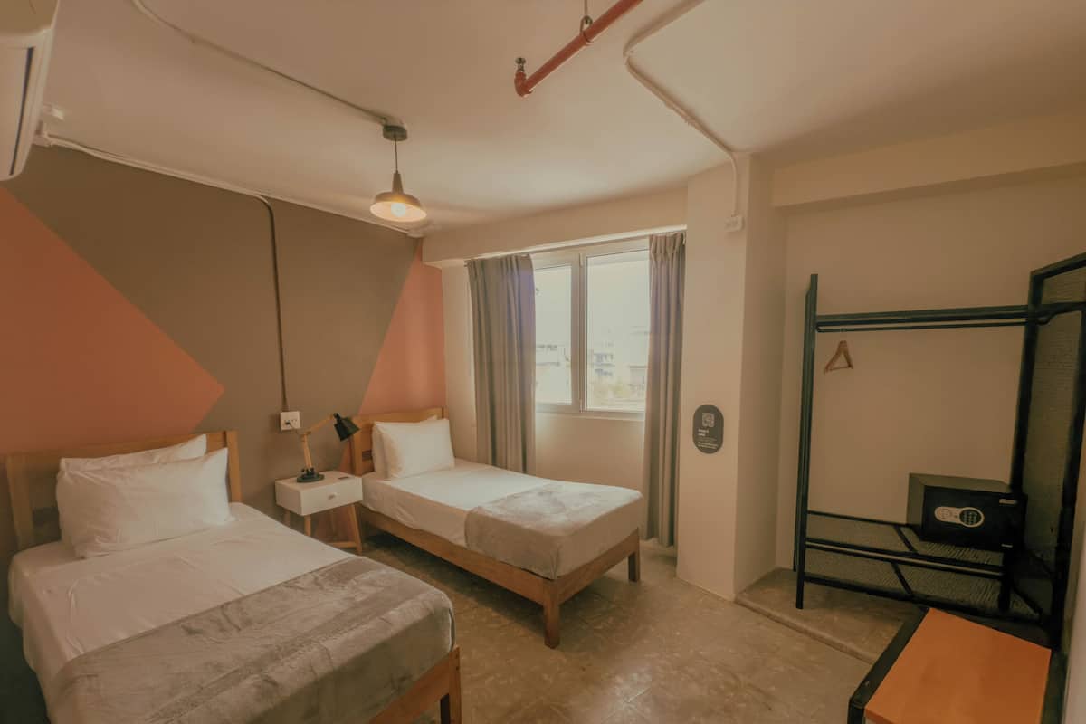Quarto de hotel com duas camas, uma escrivaninha, janela com cortinas e uma cama beliche.