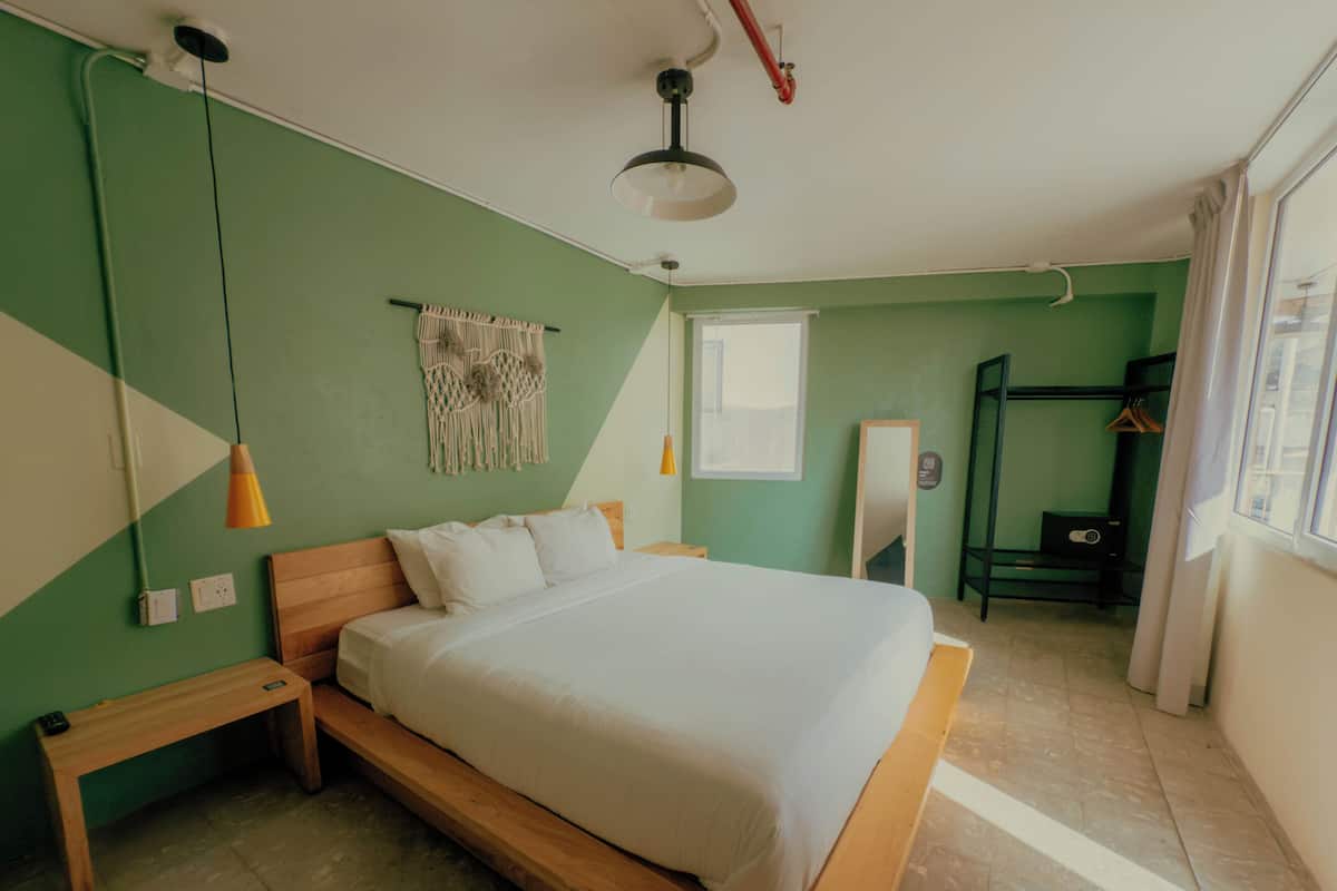 Quarto com cama, um banco, luminária pendente, tapeçaria de macramé, espelho e janela com cortinas.