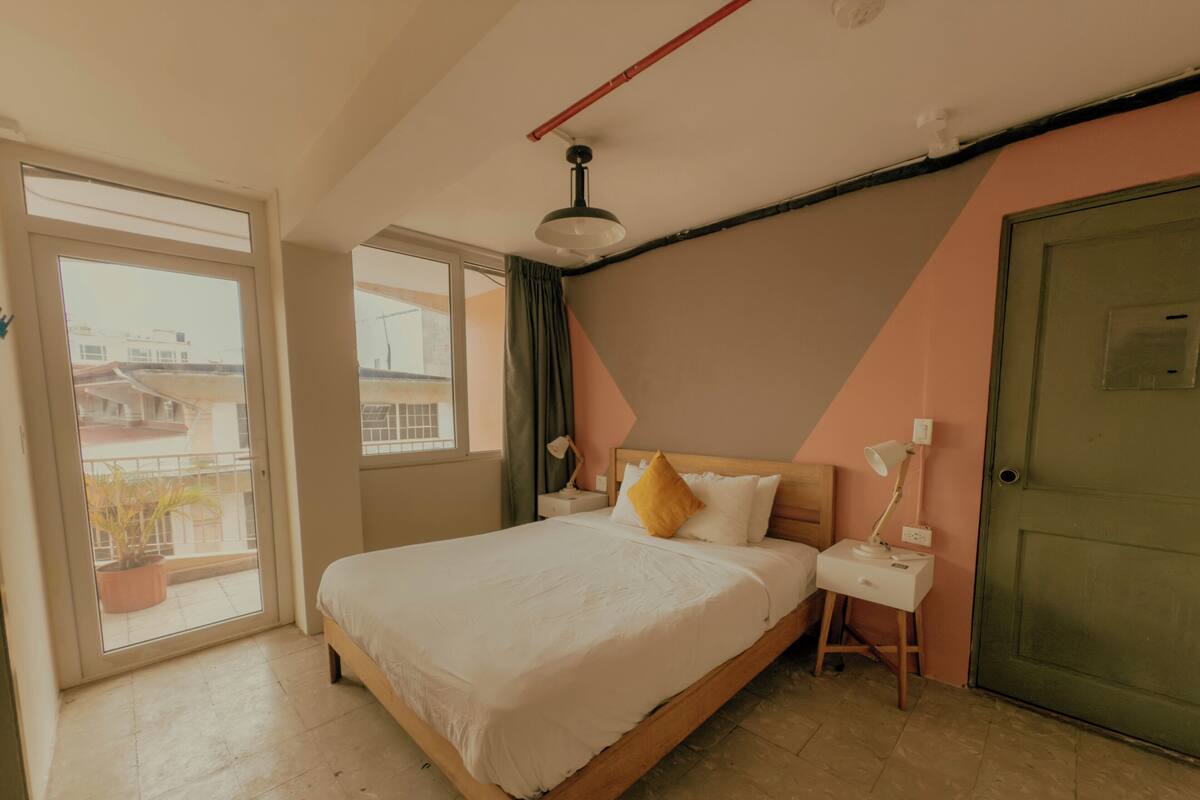 Quarto com cama, mesas de cabeceira, luminária, porta e janela.