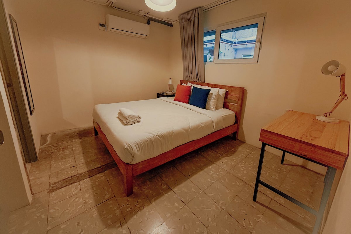 Quarto com cama, mesa de cabeceira, escrivaninha, aparelho de ar condicionado e janela com vista.