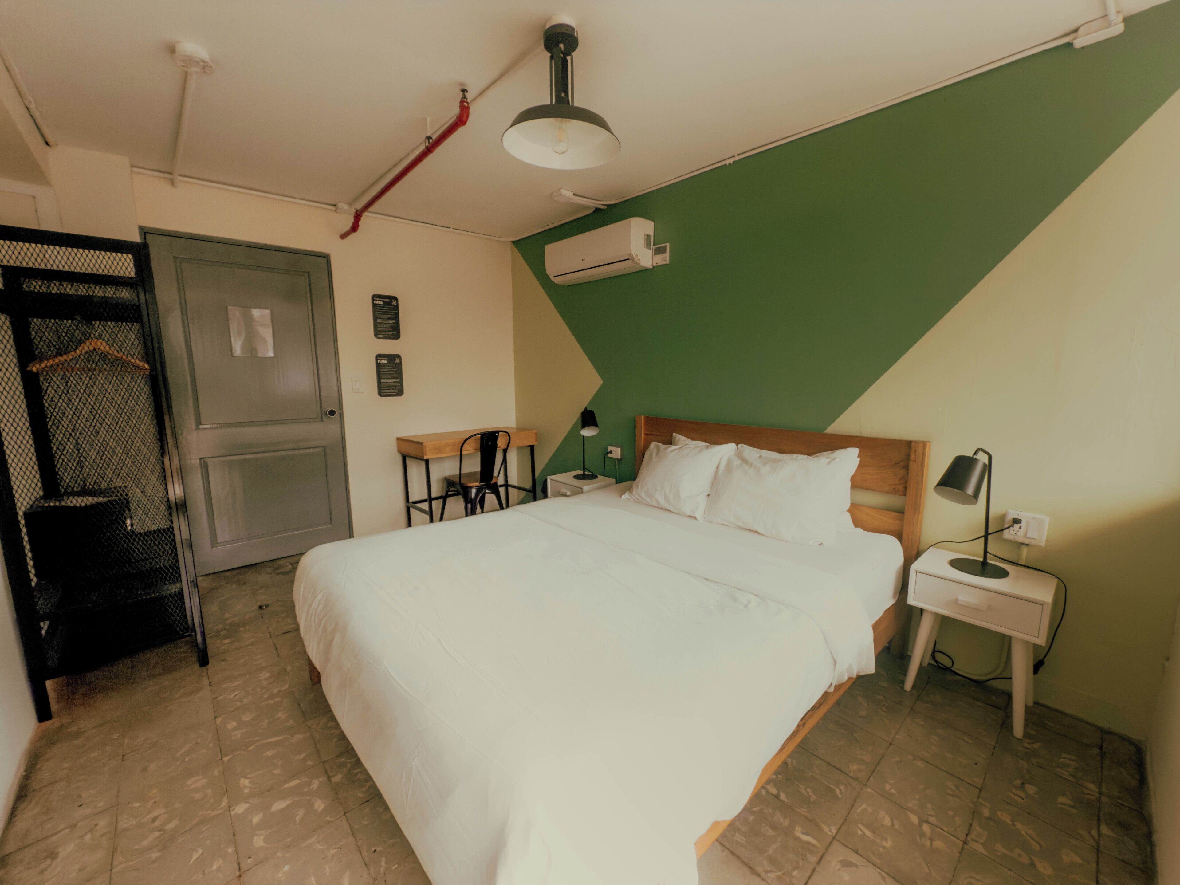 A bedroom with a bed, a desk, a chair, a nightstand, an air conditioning unit, and a door.
