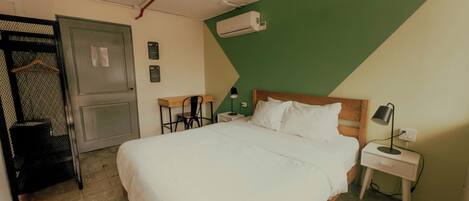 A bedroom with a bed, a desk, a chair, a nightstand, an air conditioning unit, and a door.