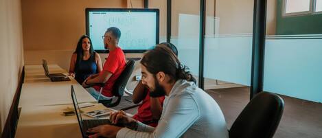 Un salón de reuniones con una pantalla grande mostrando una presentación, una computadora portátil y participantes sentados alrededor de una mesa.