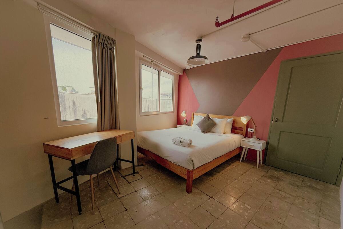 Quarto com cama, escrivaninha, cadeira, duas luminárias de apoio e janela com cortinas.