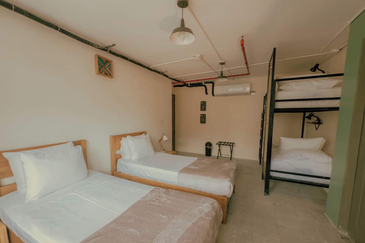 Quarto com duas camas de madeira, uma cama beliche, uma mesinha e um ar condicionado na parede.
