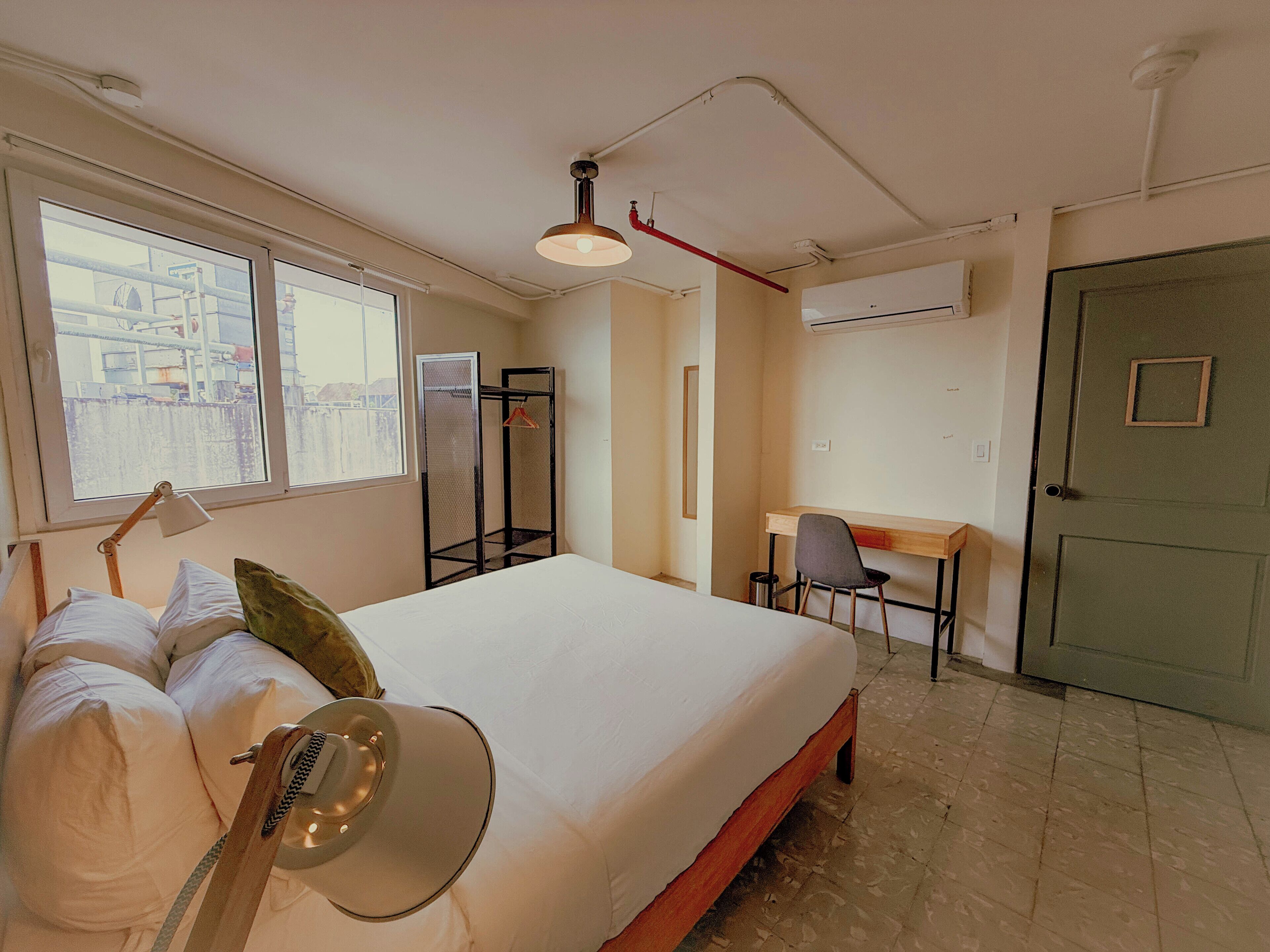 Un dormitorio con cama, escritorio, silla, guitarra y ventana con vistas a edificios.