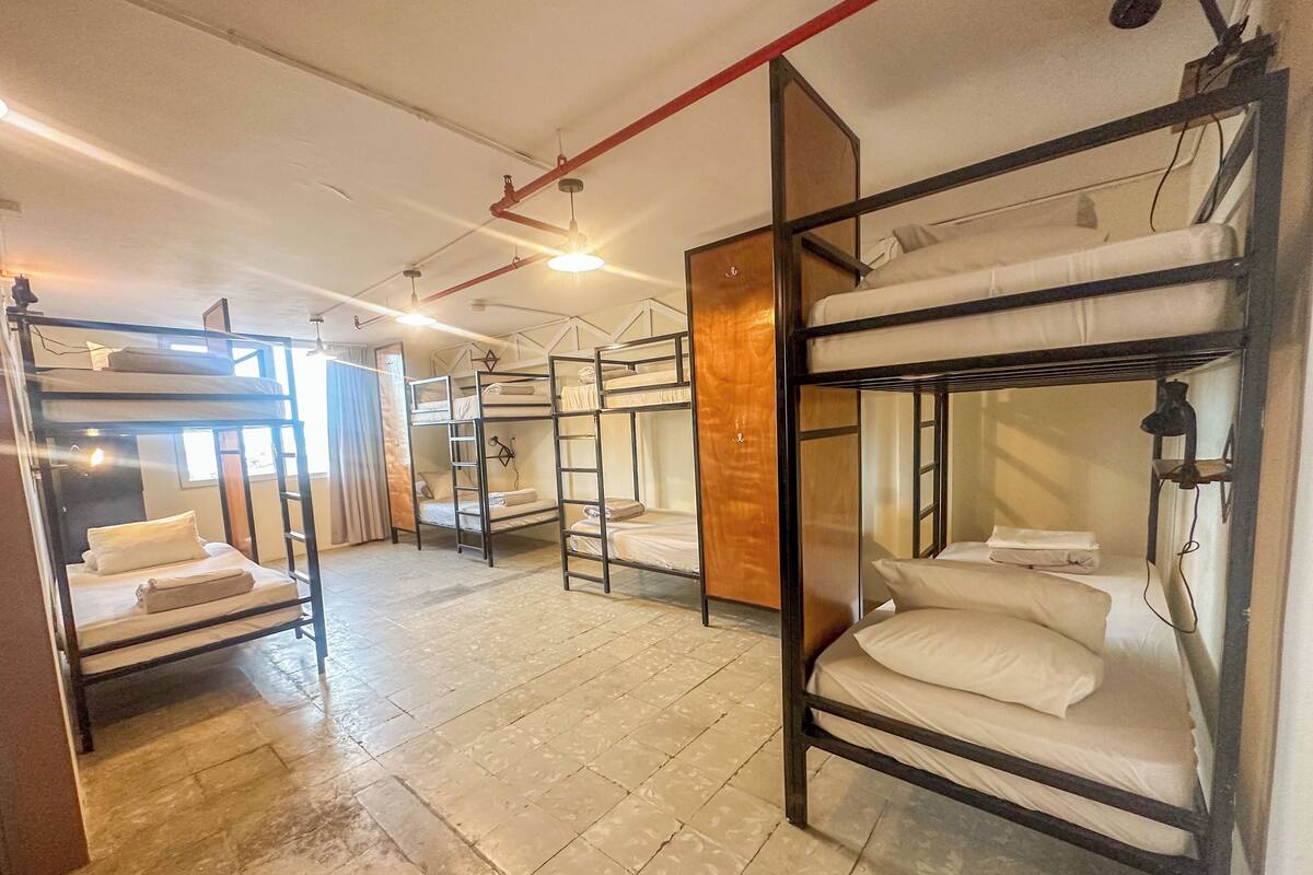 Quarto estilo dormitório com beliches, piso ladrilhado e janela com cortinas.