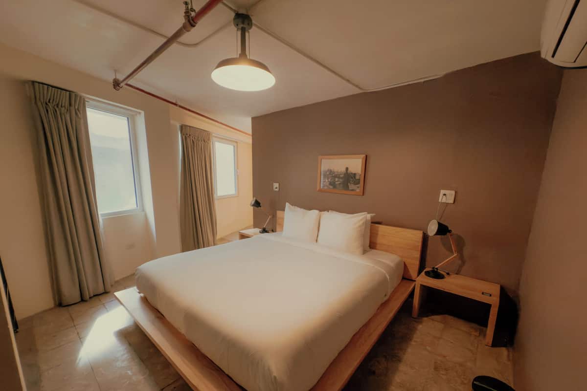 Quarto de hotel moderno com uma cama grande, mesas de cabeceira, uma luminária no teto e uma janela com cortinas.