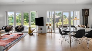 A spacious living room with a large TV, comfortable seating, a dining area, and large windows offering a view of the outdoors.