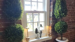 Una pared de ladrillo con una ventana, dos árboles en macetas, una mesa y una silla.