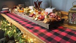 Una mesa de buffet con varios quesos, carnes y panes, sobre un mantel a cuadros, con un árbol de Navidad y luces de fondo.