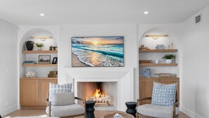 A living room with a fireplace, two armchairs, a coffee table, and a large painting of a sunset over the ocean. - Oceanview Bliss - San Clemente (San Clemente)