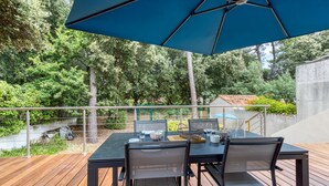 Un patio al aire libre con una mesa y sillas, una sombrilla azul y vistas de un jardín con árboles y un edificio al fondo.