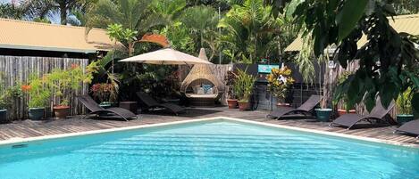 Piscina con sillas de descanso y una zona de estar a la sombra, rodeada de plantas tropicales.