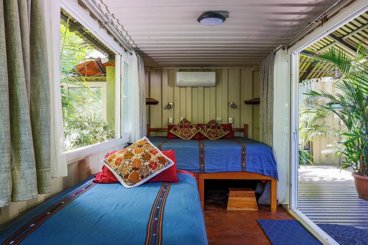 Habitación con dos camas, cada una con cubrecama azul y almohadas decorativas, un ventilador de techo y un equipo de aire acondicionado.