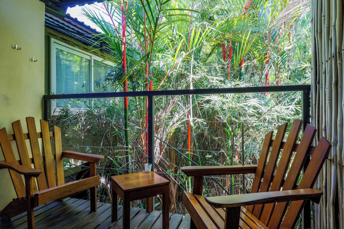 Un balcón con dos sillas de madera y una mesita, con vistas a un exuberante jardín lleno de plantas y bambú.