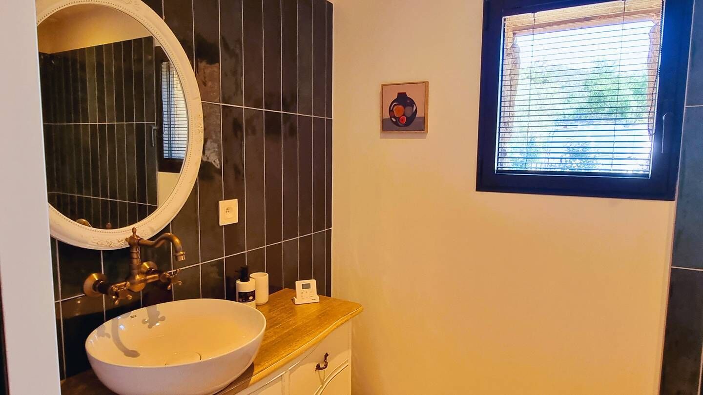 Een badkamer met een ronde spiegel, een witte wastafel, een houten wastafelmeubel, een klein raam met zonwering en een wandradiator.