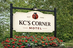 Segnale per il motel KC's Corner, con logo a forma di coccinella, fondato nel 1990, circondato da fiori colorati.