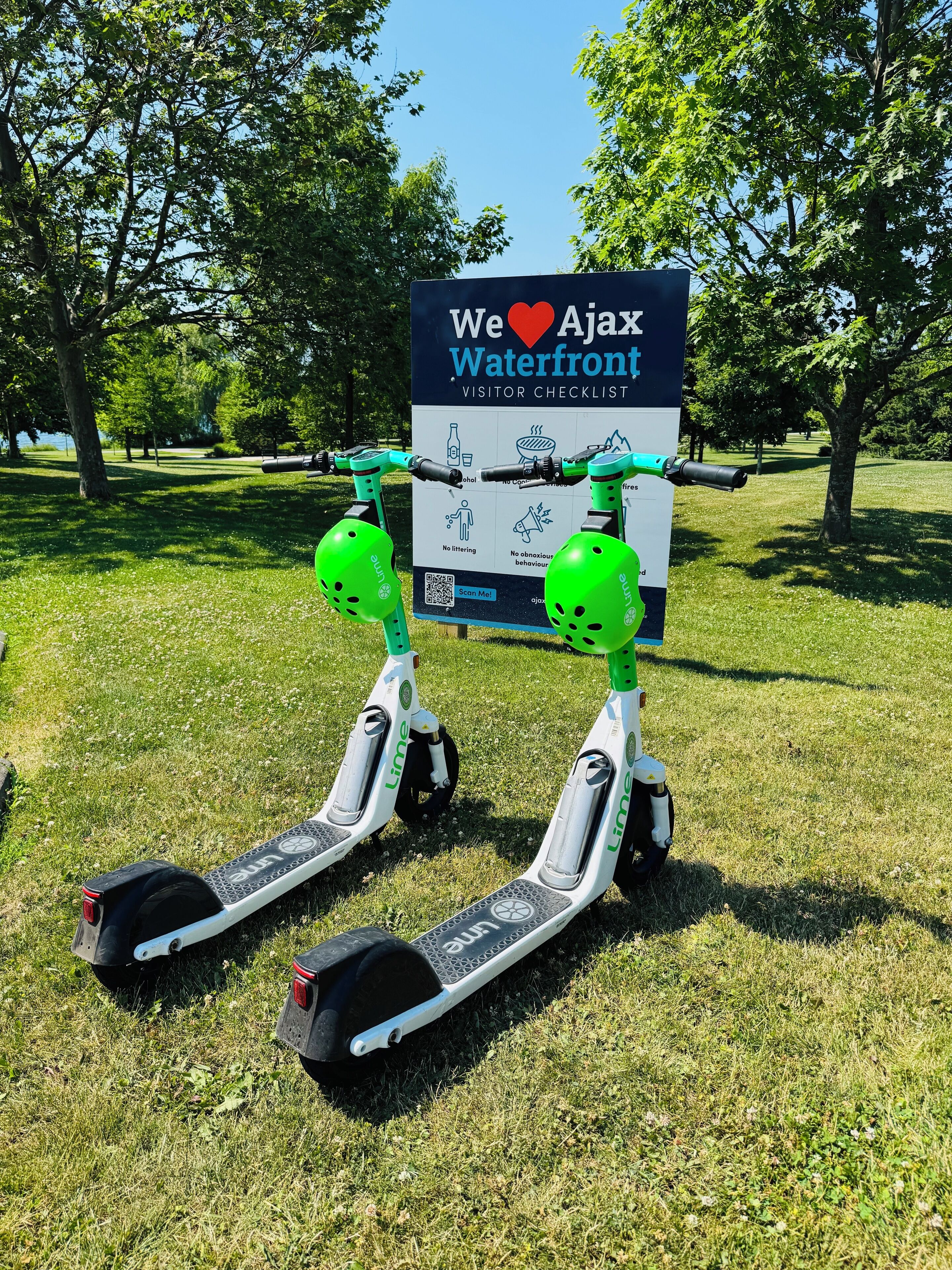 Deux trottinettes électriques Lime sont garées sur l’herbe, et on peut voir, au fond, un panneau indiquant : « Liste de contrôle pour les visiteurs du front de mer d’Ajax – Nous aimons Ajax ».
