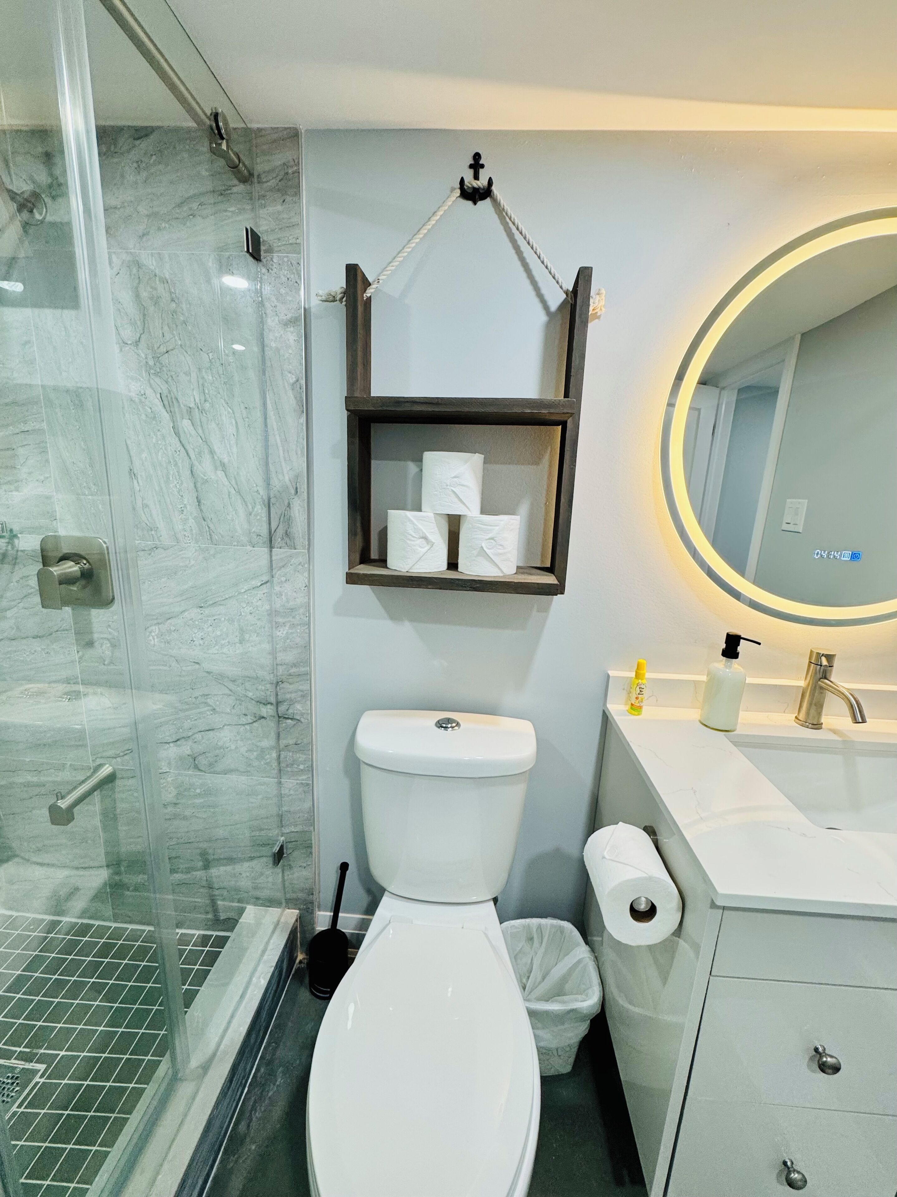 A bathroom with a glass shower enclosure, a toilet, a sink with a mirror, and a shelf with toilet paper.